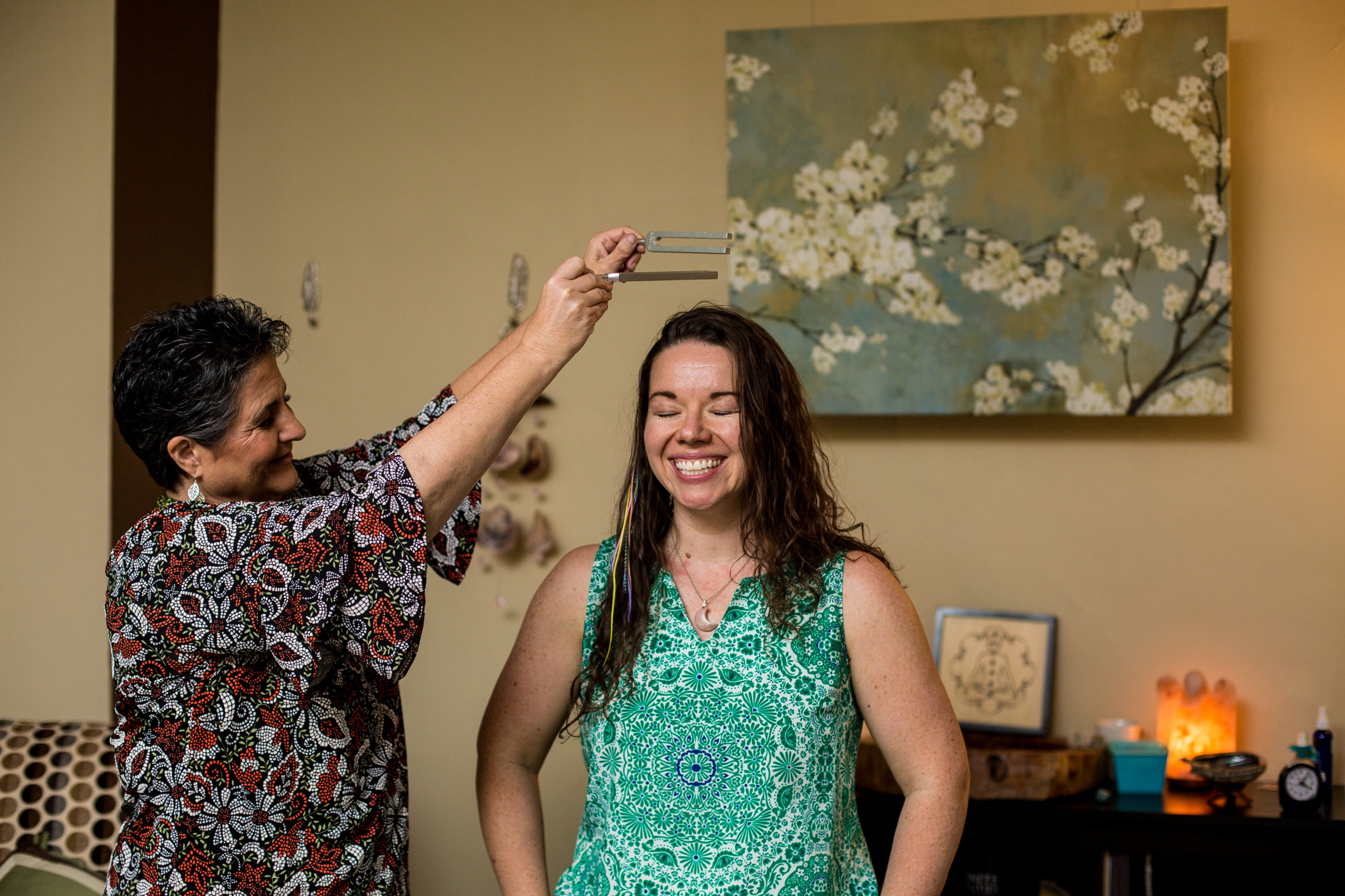Woman performs energy healing with tuning forks above another woman's head as she smiles